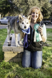 a cattle dog stands on a wooden bench bench and a white woman kneels beside him. Both are facing the camera and the woman holds a prize ribbon and is smiling.